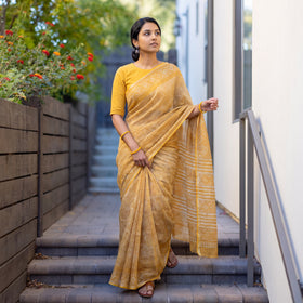 Mustard Batik Hand Block Print Kota Doria Cotton Saree, worn by a woman on garden steps beside a white wall, AI visual of the saree, iTokri