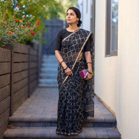 Black Batik Hand Block Print Kota Doria Cotton Saree, worn by a woman on garden steps beside a white wall, AI visual of the saree, iTokri