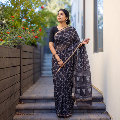Black Batik Hand Block Print Kota Doria Cotton Saree, worn by a woman on garden steps beside a white wall, AI visual of the saree, iTokri