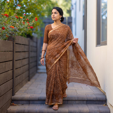 Brown Batik Hand Block Print Kota Doria Cotton Saree, worn by a woman on garden steps beside a white wall, AI visual of the saree, iTokri