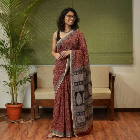 AI visual: model wearing the red natural dyed Kota Doria cotton saree with Bagh hand block print jaal and a black leaf motif pallu, in a living room