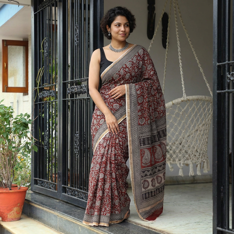 AI visual: model wearing the red natural dyed Kota Doria cotton saree with dense Bagh hand block print and a grey paisley pallu, at a doorway
