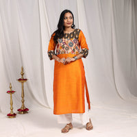 Full-length front of a bright orange art silk kurta with an olive floral yoke, brass lamps beside, festive wear